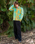 Model wearing the Kirrikin unisex Karijini long-sleeve button-down shirt featuring First Nations artwork against green foliage.