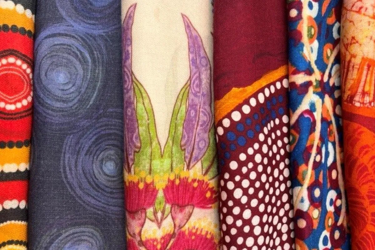 Close-up display of Kirrikin luxury silk and cashmere scarf fabrics rolled side-by-side, showcasing vibrant Aboriginal artwork motifs including floral eucalypt blossoms, dot paintings, and flowing line patterns.