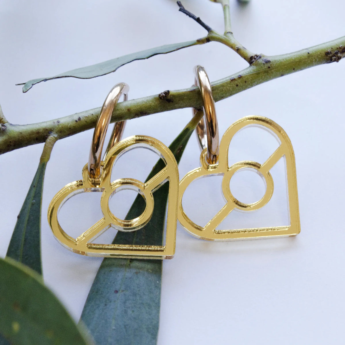 Aboriginal Gold Flag Heart Dangle Earrings hanging on a botanical branch, featuring metallic gold-toned heart cutout charms with a central ring design on gold hoop ear wires, designed by Buffie Punch.