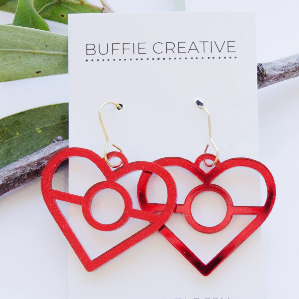 Aboriginal Red Heart Dangle Earrings on Buffie Creative card packaging, featuring cutout heart charms in reflective red with central ring motifs on gold-toned ear wires, designed by Buffie Punch.