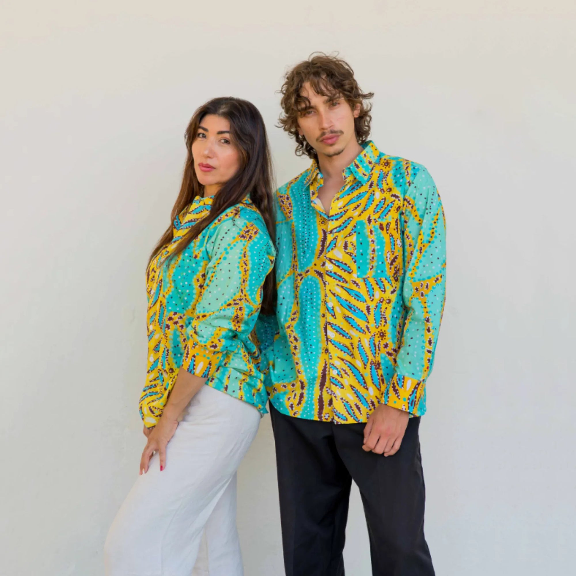 Male and female models wearing matching Kirrikin unisex Karijini long-sleeve shirts featuring vibrant turquoise and yellow Indigenous artwork by Sally White.