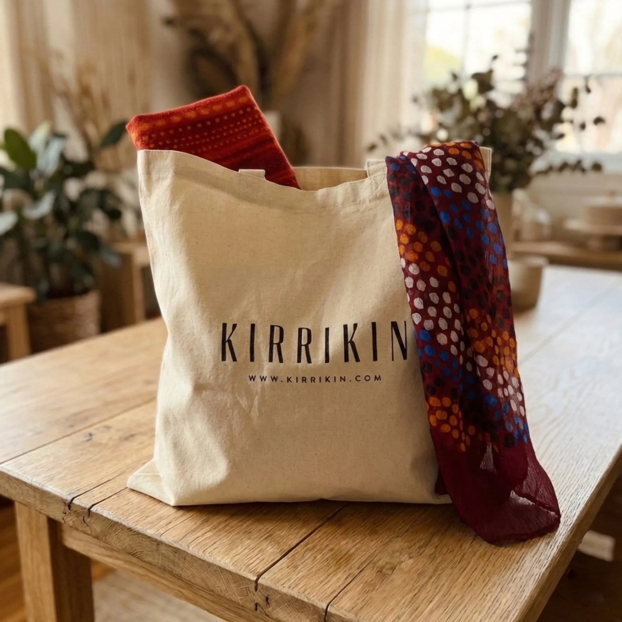 A beige canvas tote bag with the brand name 'KIRRIKIN' printed in black, sitting on a rustic wooden table. Two patterned scarves are draped from the top of the bag, one in rust red and orange, and the other in deep burgundy with orange, blue, and white geometric patterns.