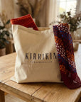 A beige canvas tote bag with the brand name 'KIRRIKIN' printed in black, sitting on a rustic wooden table. Two patterned scarves are draped from the top of the bag, one in rust red and orange, and the other in deep burgundy with orange, blue, and white geometric patterns.
