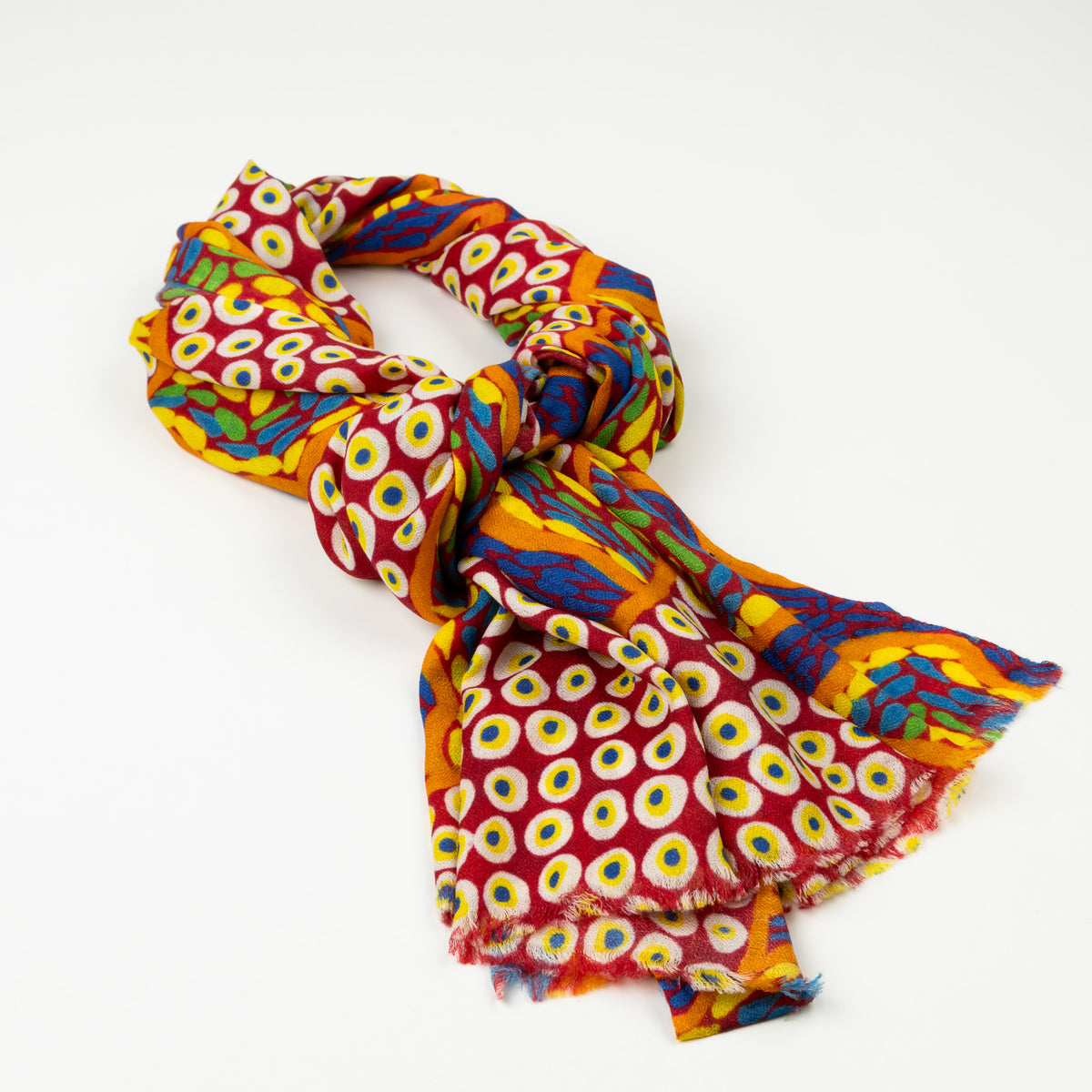 Kirrikin Stories In The Sun Luxury Cashmere Scarf styled in a loose knot on a white background, featuring red, yellow, and blue Indigenous circle artwork by Kaye White with frayed edges.