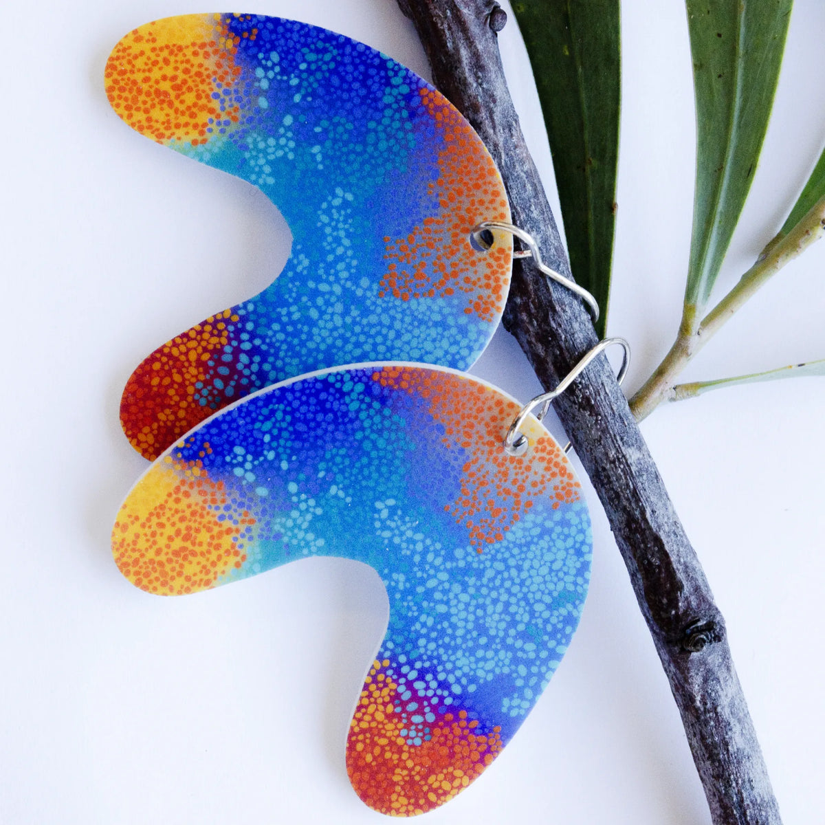 Large U Shape Makuru Earrings hanging against a botanical branch background, featuring curved U-shaped charms with vivid blue, purple, and orange Indigenous dot artwork on silver-toned ear wires, designed by Buffie Punch.