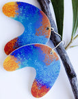Large U Shape Makuru Earrings hanging against a botanical branch background, featuring curved U-shaped charms with vivid blue, purple, and orange Indigenous dot artwork on silver-toned ear wires, designed by Buffie Punch.